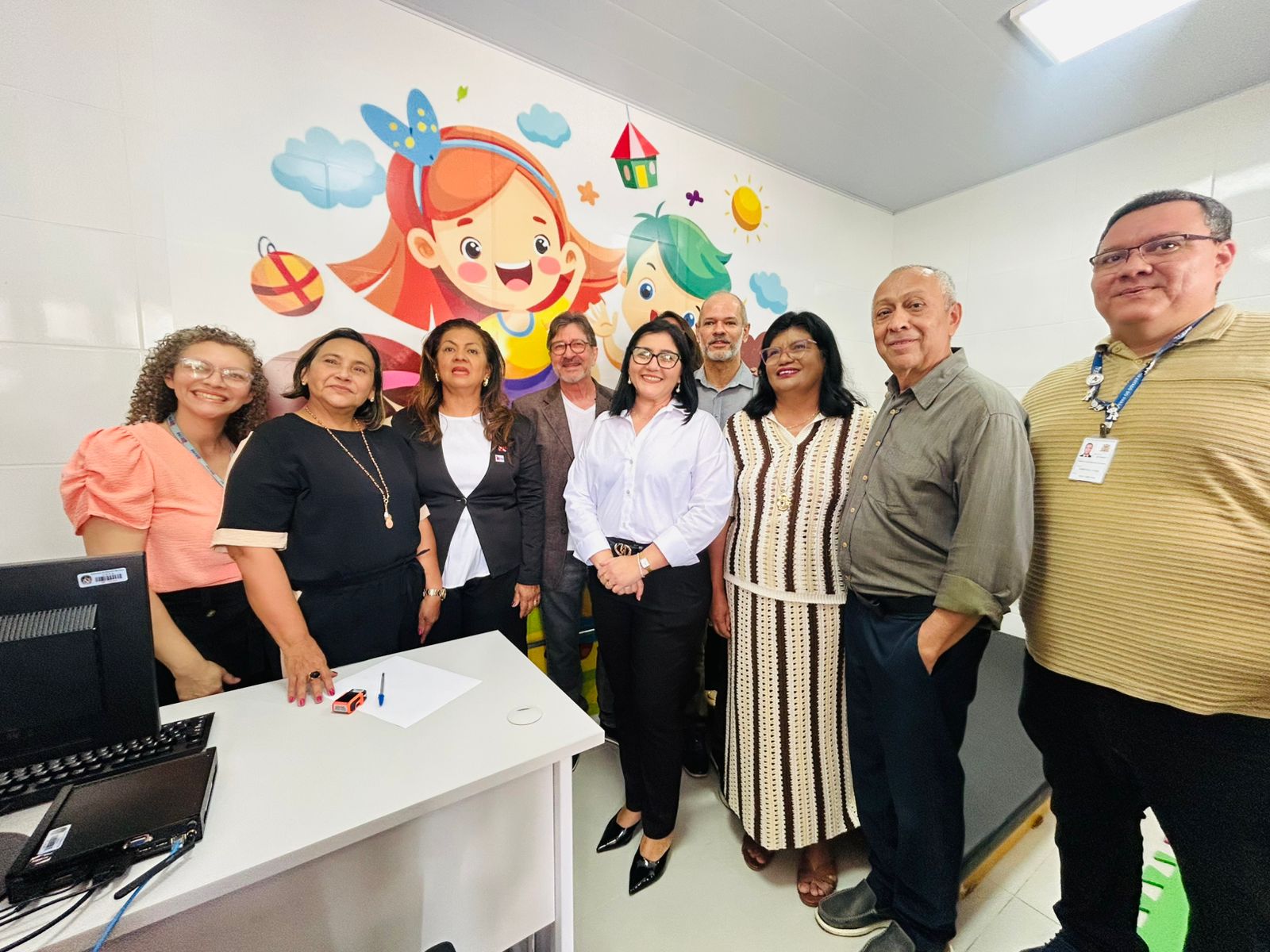 Um grupo de dez pessoas posa para uma foto em uma sala clara, com uma parede decorada por um grande mural infantil colorido, com desenhos de uma menina sorridente, brinquedos e elementos como sol e nuvens. As pessoas estão alinhadas lado a lado, olhando para a câmera e sorrindo. À esquerda há uma mesa com computador e documentos.