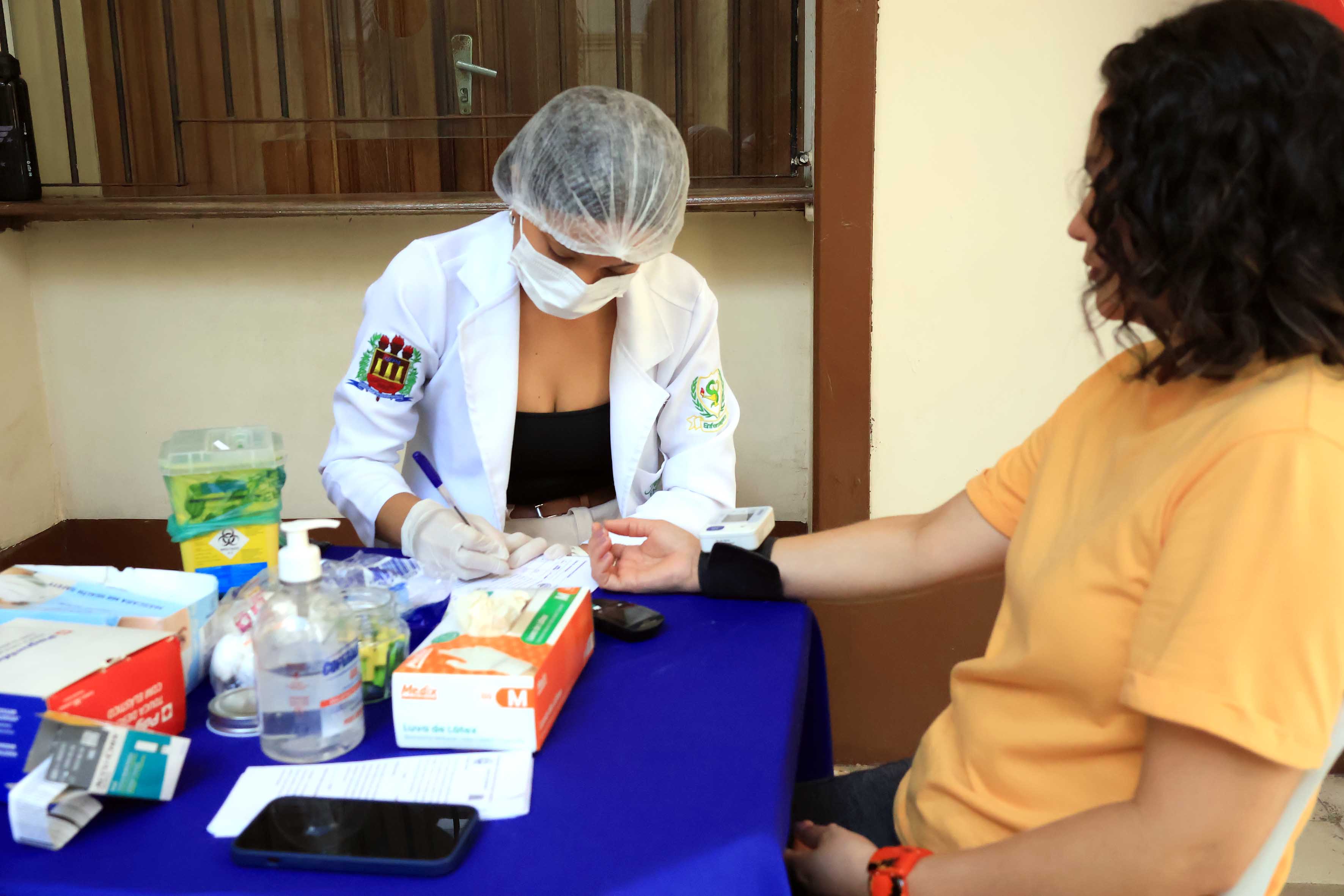 A imagem mostra uma profissional de saúde, usando jaleco branco, luvas e máscara, que realiza um procedimento em uma paciente sentada.  Concentrada, a profissional, inclinada sobre uma mesa azul com insumos de saúde, anota dados enquanto coleta material da paciente à direita vestindo uma camiseta laranja, com o braço exposto para o procedimento. A cena capturada durante uma campanha de prevenção, reflete um ambiente de cuidado e saúde pública. O fundo mostra uma janela de madeira e parede clara.