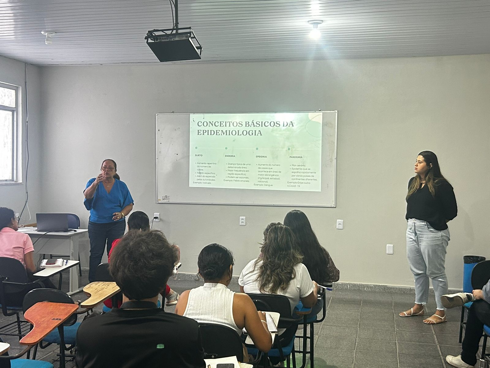 A imagem mostra uma sala de aula com uma apresentação em andamento. Há uma tela de projetor exibindo um slide intitulado “CONCEITOS BÁSICOS DA EPIDEMIOLOGIA”. A sala tem paredes brancas e há estudantes sentados de frente para a tela, alguns fazendo anotações. Duas pessoas estão conduzindo a aula; uma está em pé perto da tela apontando para ela, e a outra está em pé no lado direito da imagem. 