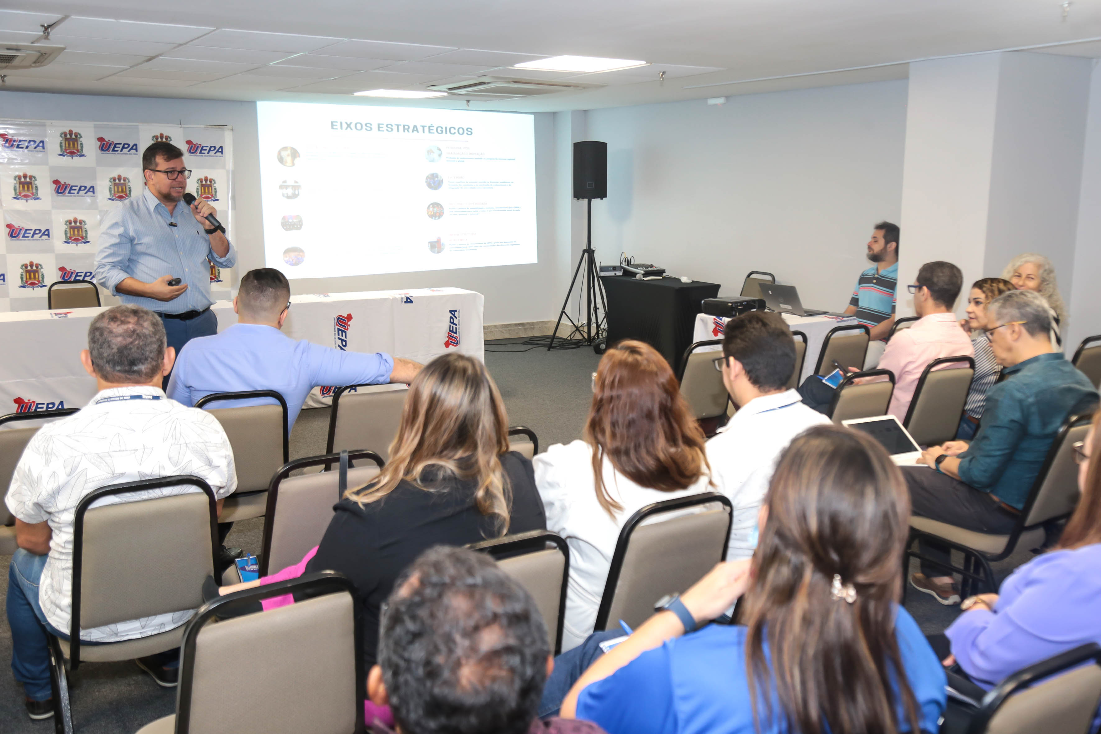 A imagem retrata um ambiente interno onde está ocorrendo uma apresentação. Na frente, há fileiras de pessoas sentadas, de frente para a parte da frente, onde um apresentador está à esquerda de uma tela de projeção. A plateia parece estar ouvindo atentamente. A tela de projeção exibe texto e gráficos, mas o conteúdo específico não é claramente visível devido à resolução e ao ângulo da fotografia. A sala tem iluminação fluorescente e parece estar preparada para um evento profissional ou conferência. 