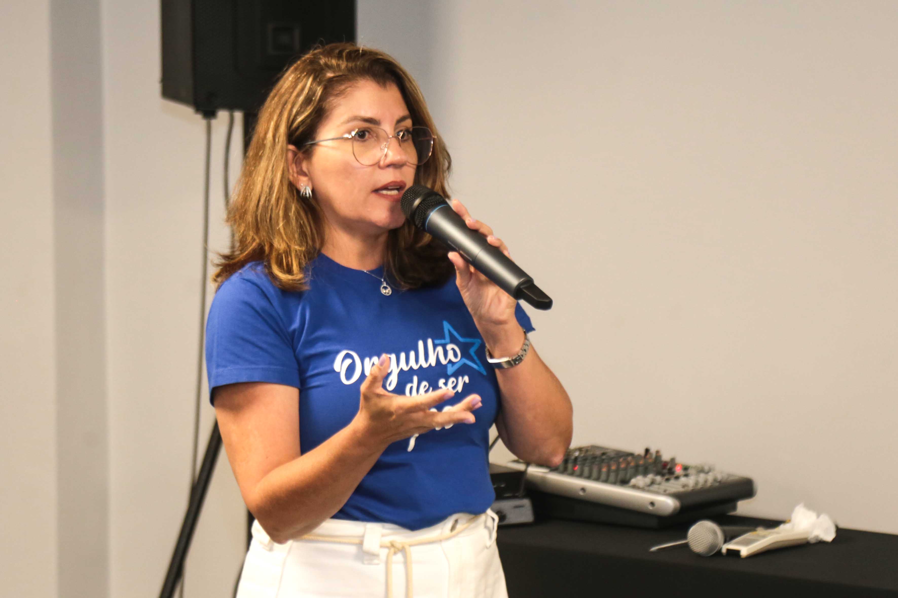 A imagem retrata uma pessoa em pé na frente de um microfone, aparentando estar falando ou apresentando. A pessoa está vestindo uma camiseta azul com texto branco, calças brancas e tem um relógio de pulso no pulso esquerdo. Ao fundo, há equipamentos de áudio, incluindo o que parece ser uma mesa de som e cabos. 