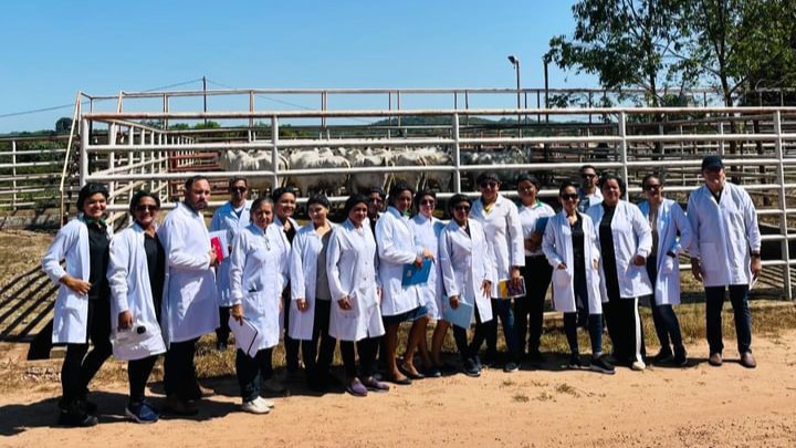A imagem mostra um grupo de alunos em frente a um conjunto de currais para animais.  Cada um deles usa jaleco branco, pois são estudantes do curso de Engenharia de Alimentos.O cenário de fundo apresenta estruturas ao ar livre típicas de uma fazenda ou instalação de pesquisa agrícola. 