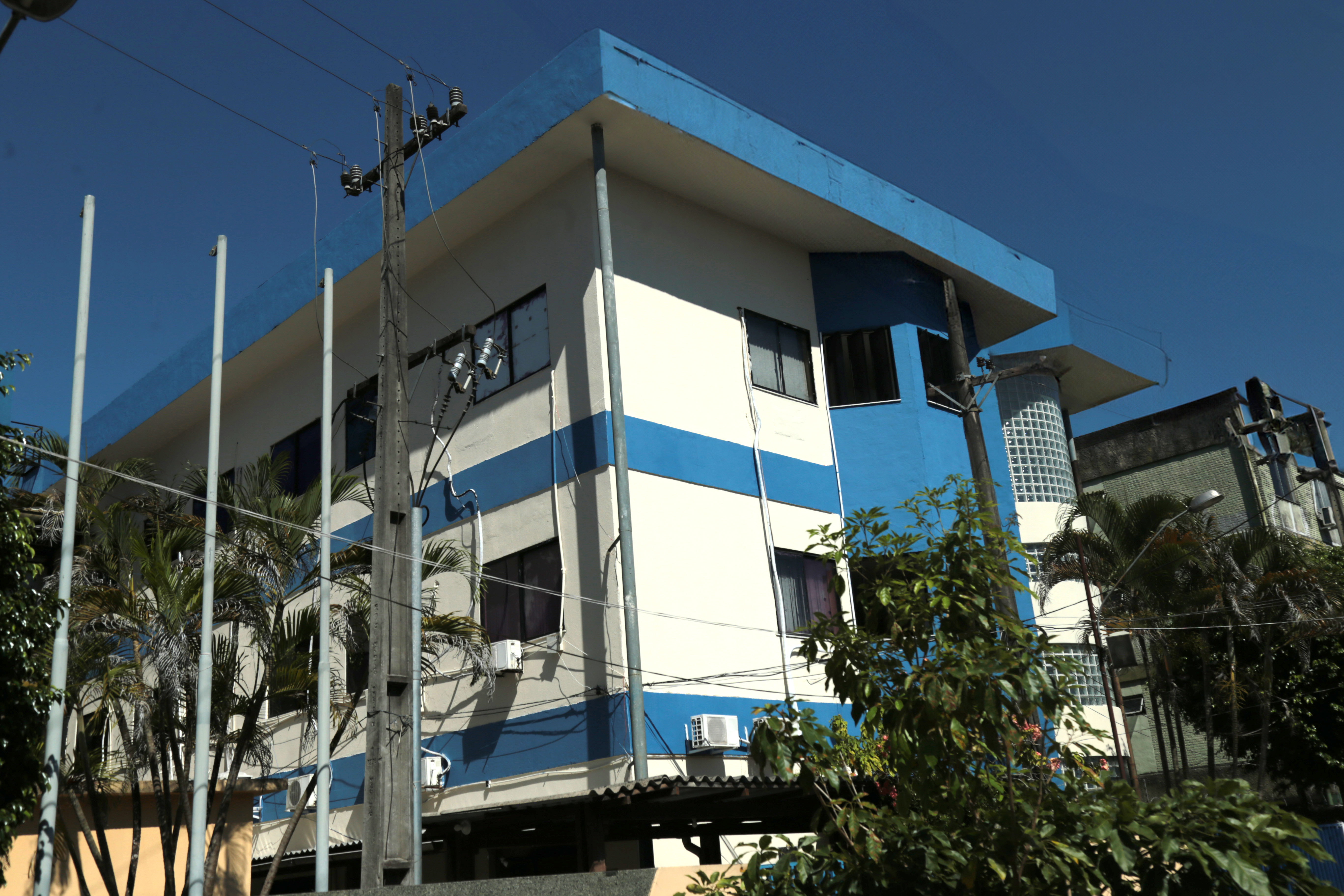 A imagem mostra um prédio de fachada moderna, com tons de azul e branco, possivelmente uma instituição ou edifício comercial. As janelas são amplas, com vidro refletindo o céu. À frente, há uma área verde com grama e palmeiras. Uma pessoa está entrando no prédio, carregando uma mochila. O céu está limpo, indicando um dia ensolarado. Outro edifício similar está ao fundo, também com pintura azul e branca.