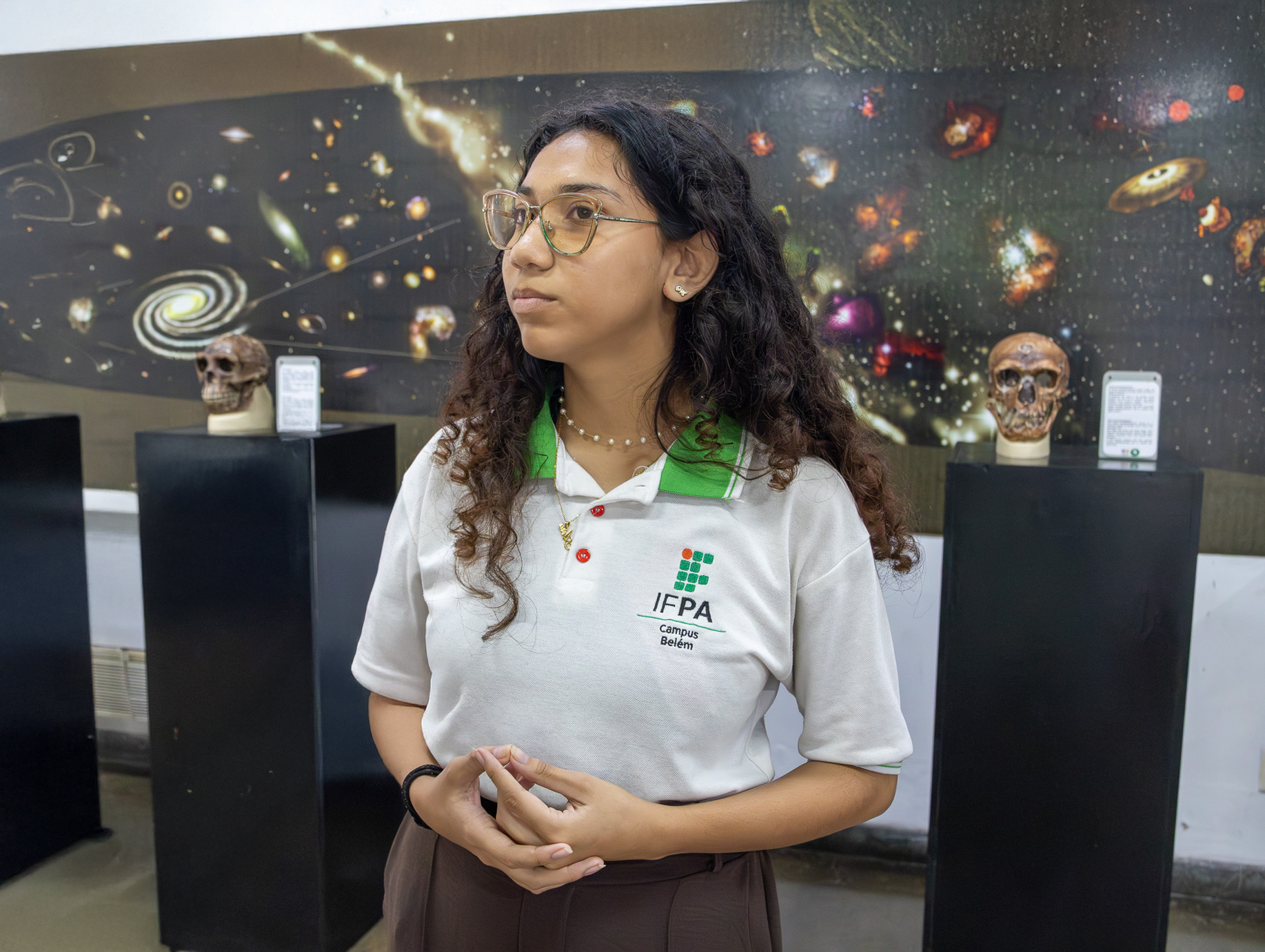 Uma jovem com uniforme escolar é entrevistada diante de um mural espacial. Ao fundo, há modelos de crânios em pedestais e placas informativas, compondo um ambiente de exposição científica