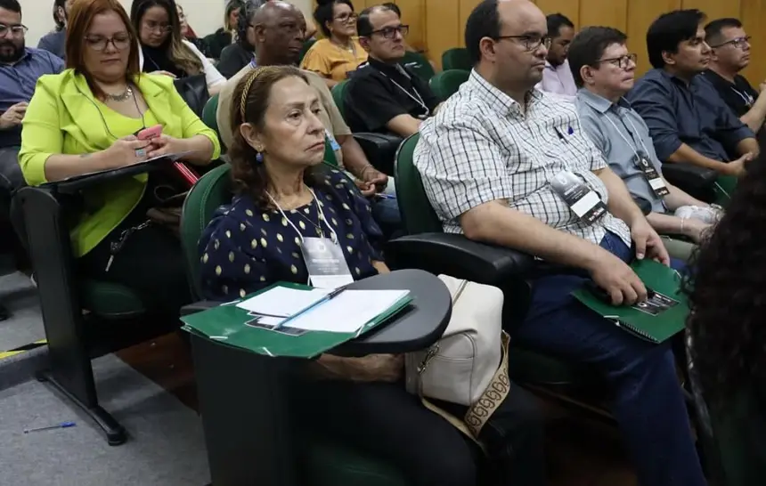 A imagem mostra um grupo de pessoas sentadas no auditório, participando de um evento. Elas estão atentas às atividades, com pastas e crachás de identificação sobre as mesas individuais. O ambiente tem clima formal, típico de encontros acadêmicos ou institucionais.