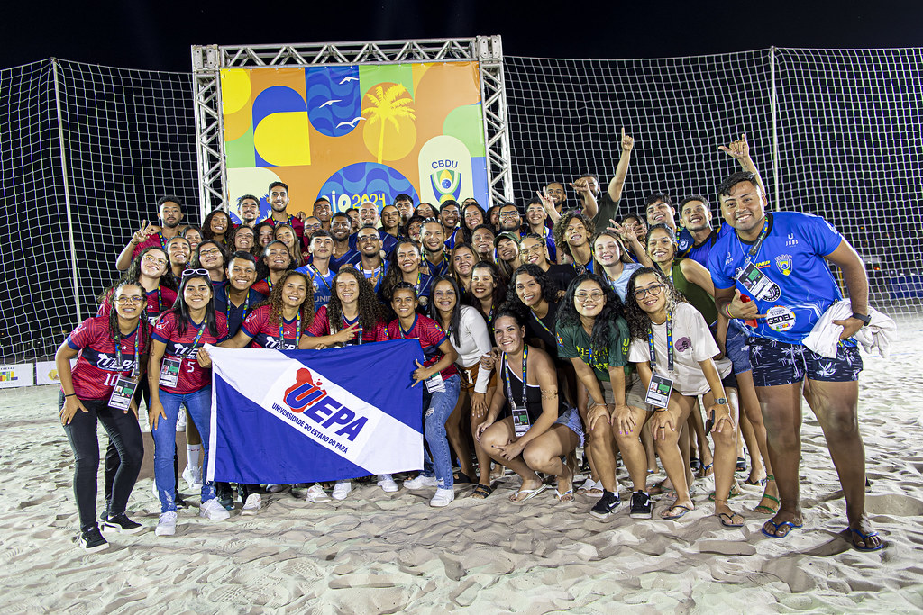 A imagem mostra um grande grupo de estudantes, todos com sorrisos no rosto, reunidos em uma praia à noite. Eles estão celebrando e posando para a foto enquanto seguram uma bandeira da Universidade do Estado do Pará (Uepa). A bandeira tem as cores azul, branco e vermelho, com o nome da universidade claramente visível. Ao fundo, é possível ver uma grande estrutura metálica que sustenta um painel colorido com o emblema da Confederação Brasileira do Desporto Universitário (CBDU).