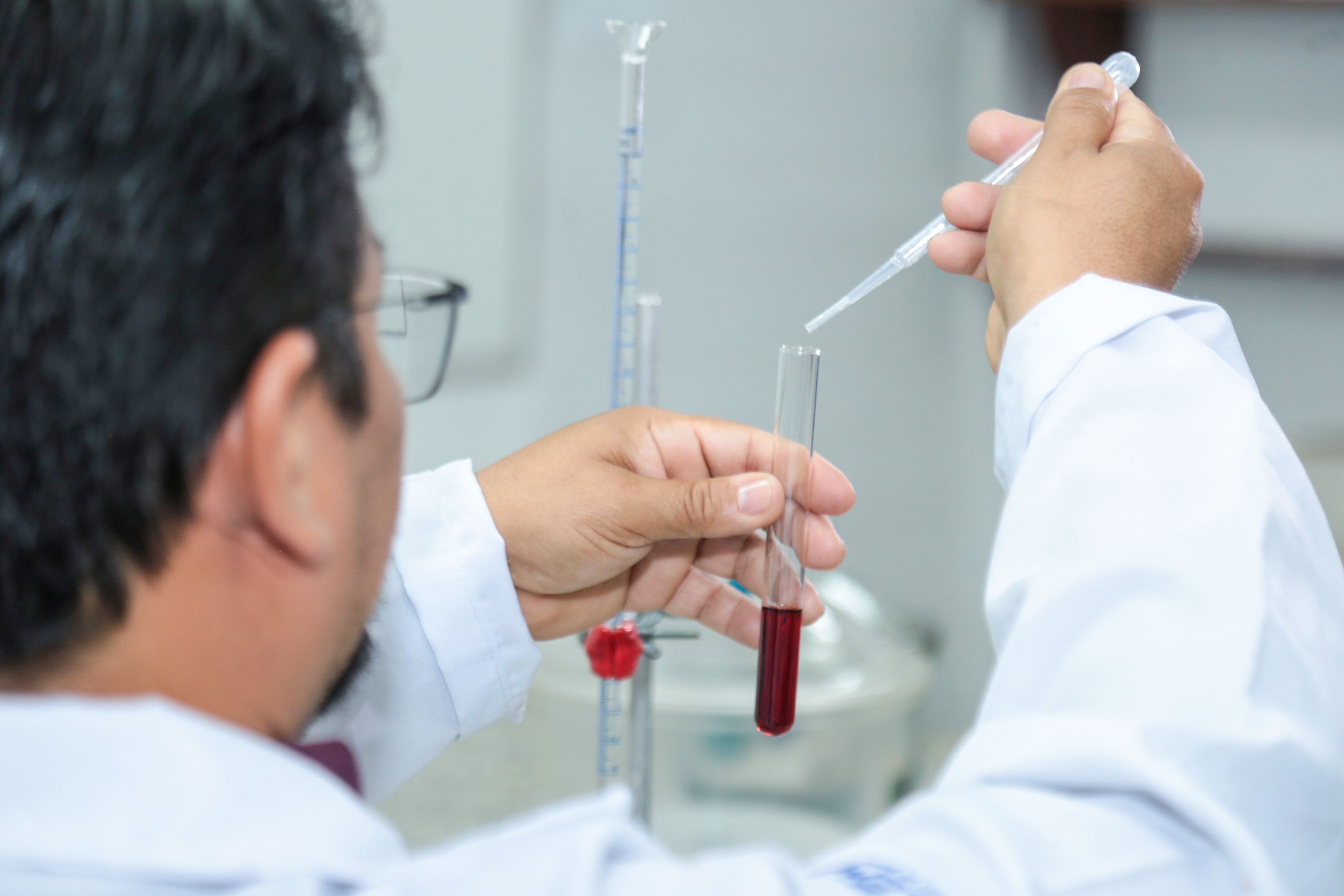 Na imagem, um homem, parcialmente de costas e vestindo jaleco branco, está manipulando equipamentos de laboratório. Ele segura um tubo de ensaio transparente com um líquido vermelho, enquanto utiliza uma pipeta para adicionar ou retirar o líquido. Ao fundo, há outros instrumentos de laboratório levemente desfocados, como uma bureta presa em um suporte vertical.