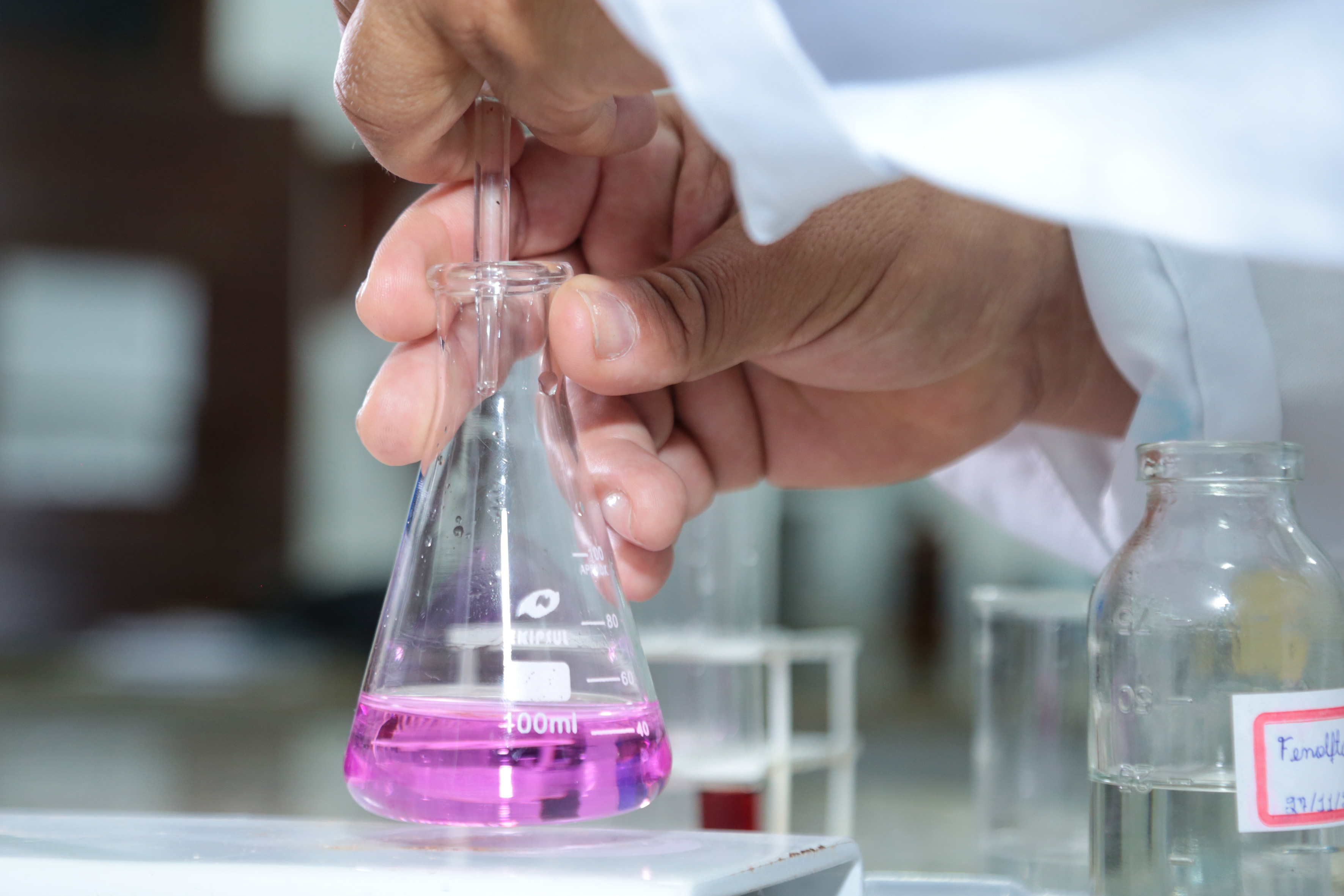 A imagem mostra uma pessoa manuseando um erlenmeyer com líquido rosa em um laboratório de química, utilizando uma pipeta. A pessoa veste jaleco branco, indicando uma atividade científica ou experimental.