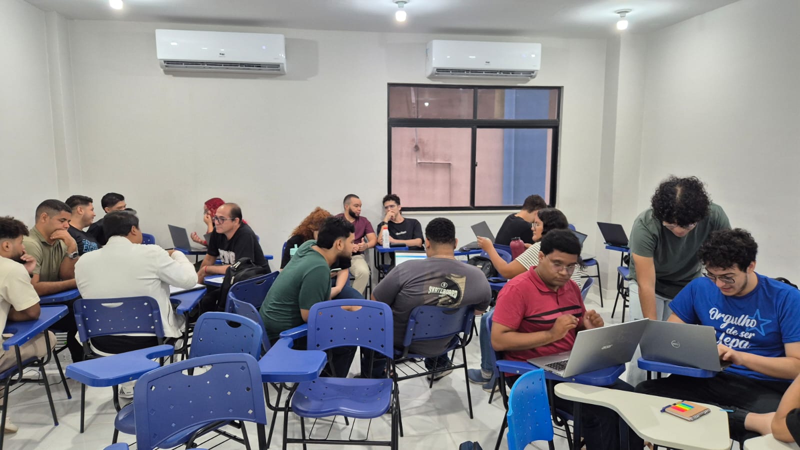 A imagem mostra uma sala de aula com grupos de estudantes reunidos em atividades colaborativas. As carteiras estão organizadas de forma a facilitar a interação, e vários alunos utilizam notebooks para desenvolver seus trabalhos.