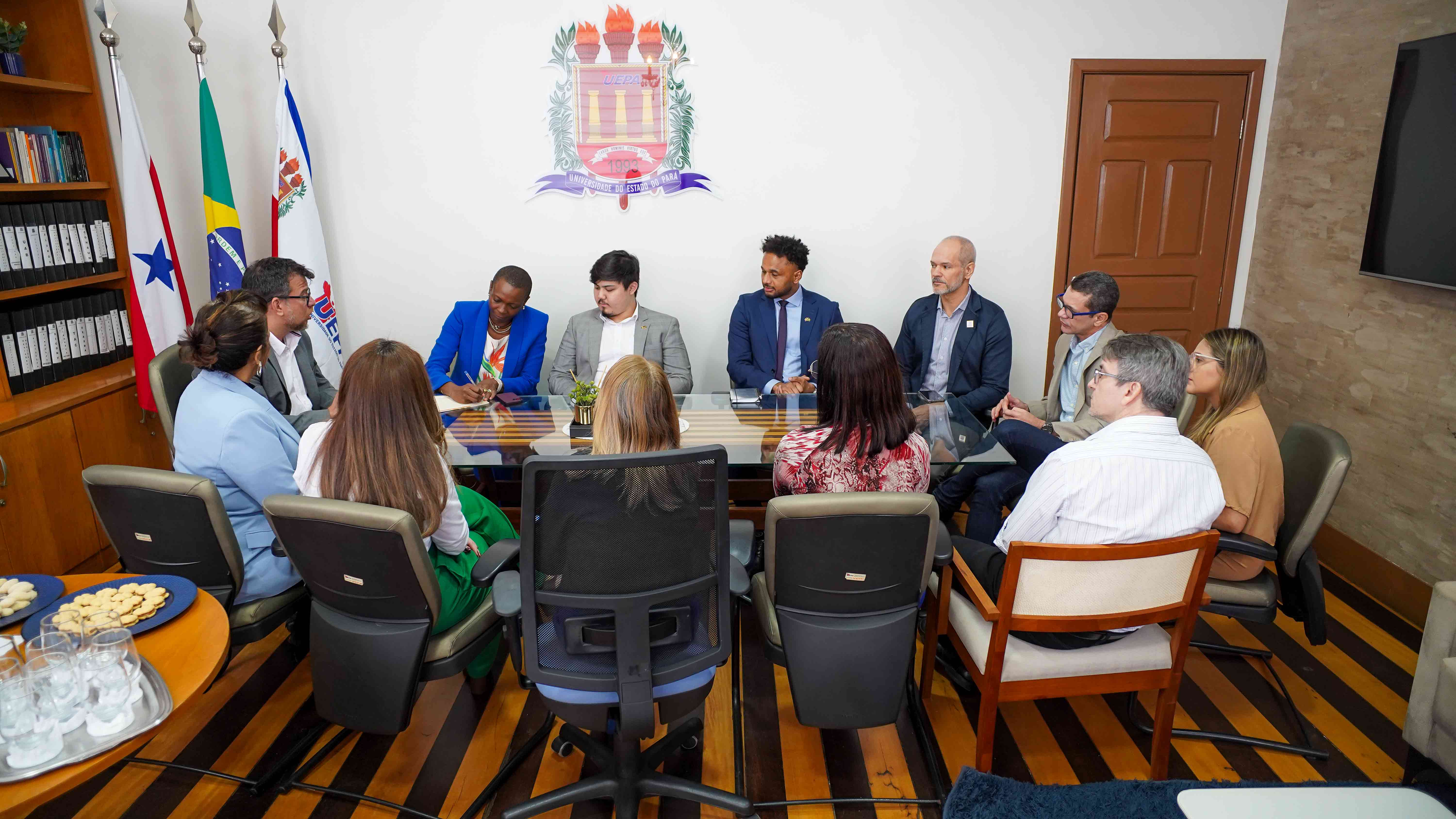 A imagem retrata um grupo de pessoas sentadas ao redor de uma mesa retangular comprida em uma sala de reuniões. O ambiente possui um carpete azul e paredes brancas decoradas com bandeiras e quadros. Uma grande janela permite a entrada de luz natural. Os indivíduos estão vestidos com trajes formais, sugerindo uma reunião ou conferência. Documentos, laptops e garrafas de água estão sobre a mesa, indicando uma discussão ativa ou sessão de trabalho.