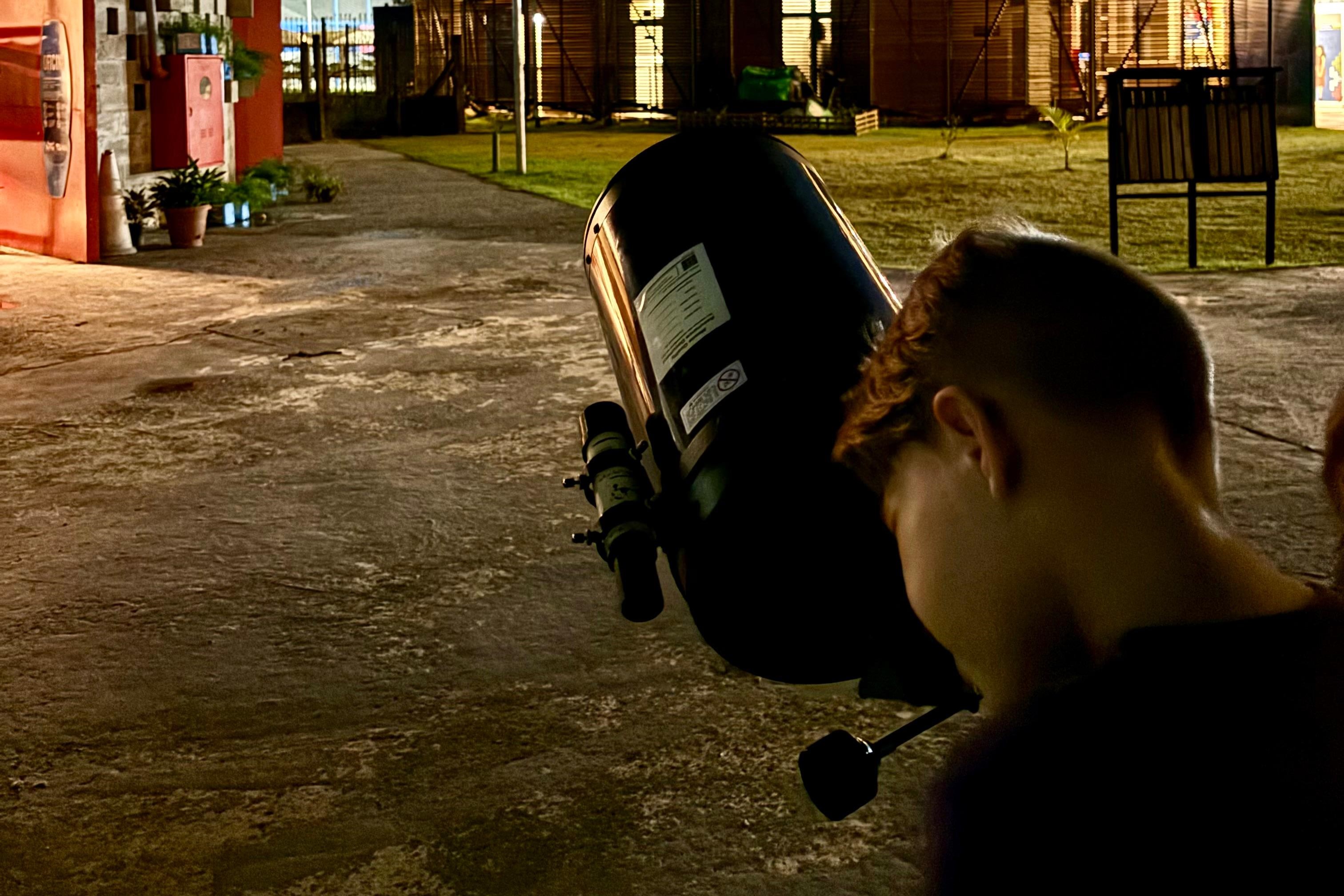 A imagem mostra uma pessoa, de costa para a câmera, utilizando um telescópio para a observação do céu notuno