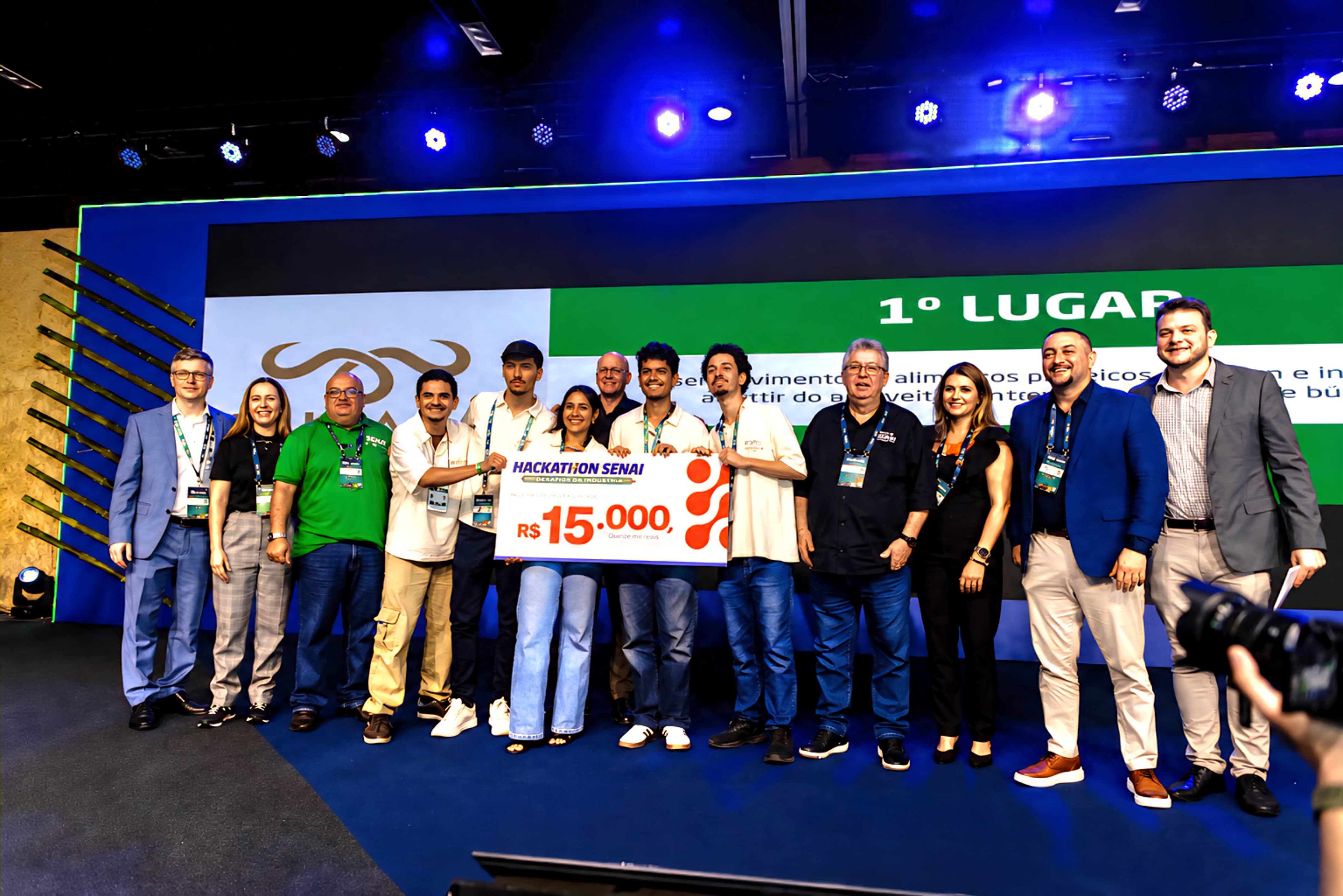 Grupo de estudantes e representantes de instituições posa no palco durante uma cerimônia de premiação, segurando um cheque simbólico de R$ 15 mil do Hackathon SENAI. Ao fundo, um telão destaca a conquista do 1º lugar, enquanto a equipe sorri para a foto oficial sob iluminação cênica azul.