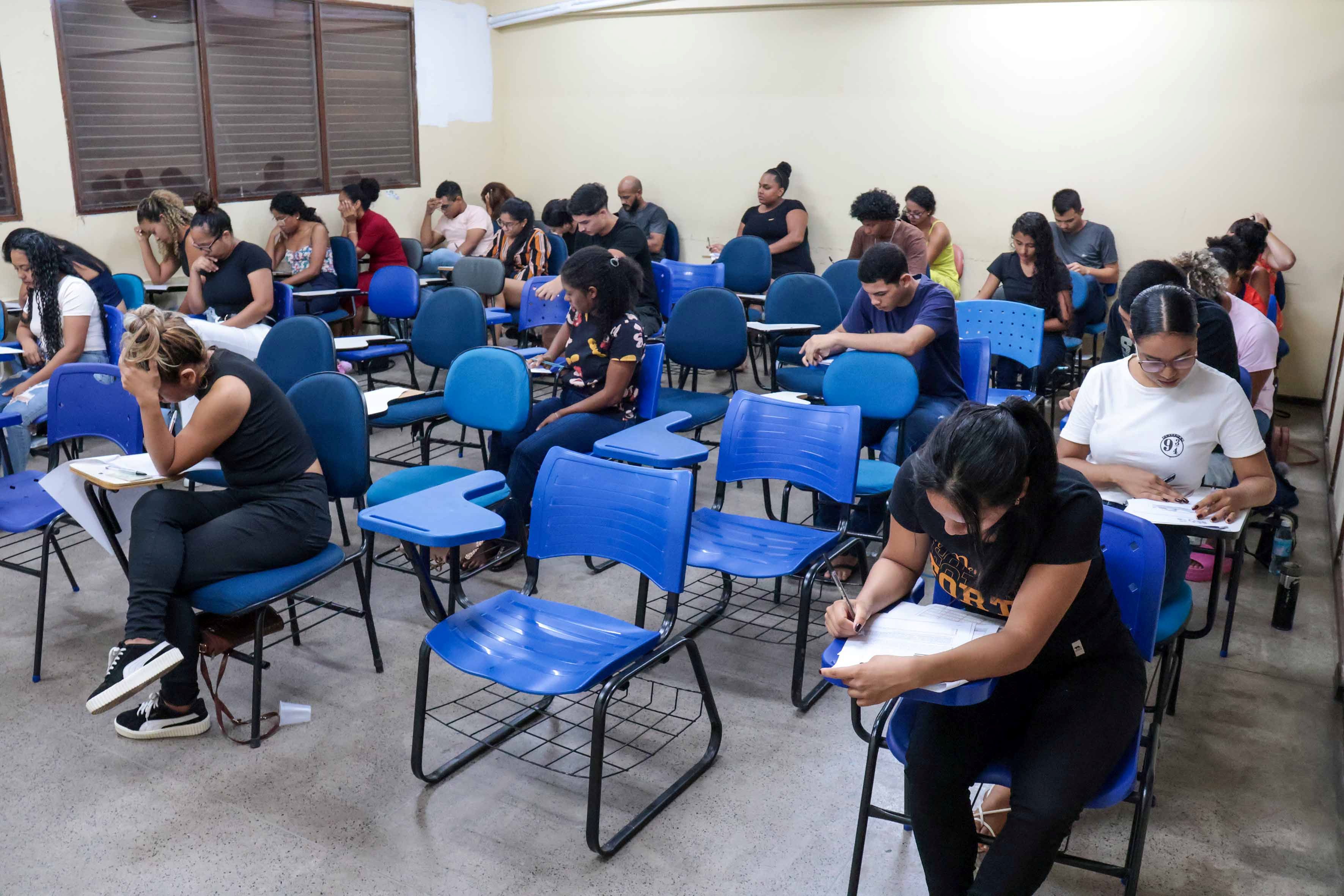 A foto mostra uma sala de aula com várias pessoas sentadas em carteiras escolares, concentradas enquanto realizam uma prova ou preenchendo formulários. O ambiente é simples, com cadeiras azuis e paredes claras.