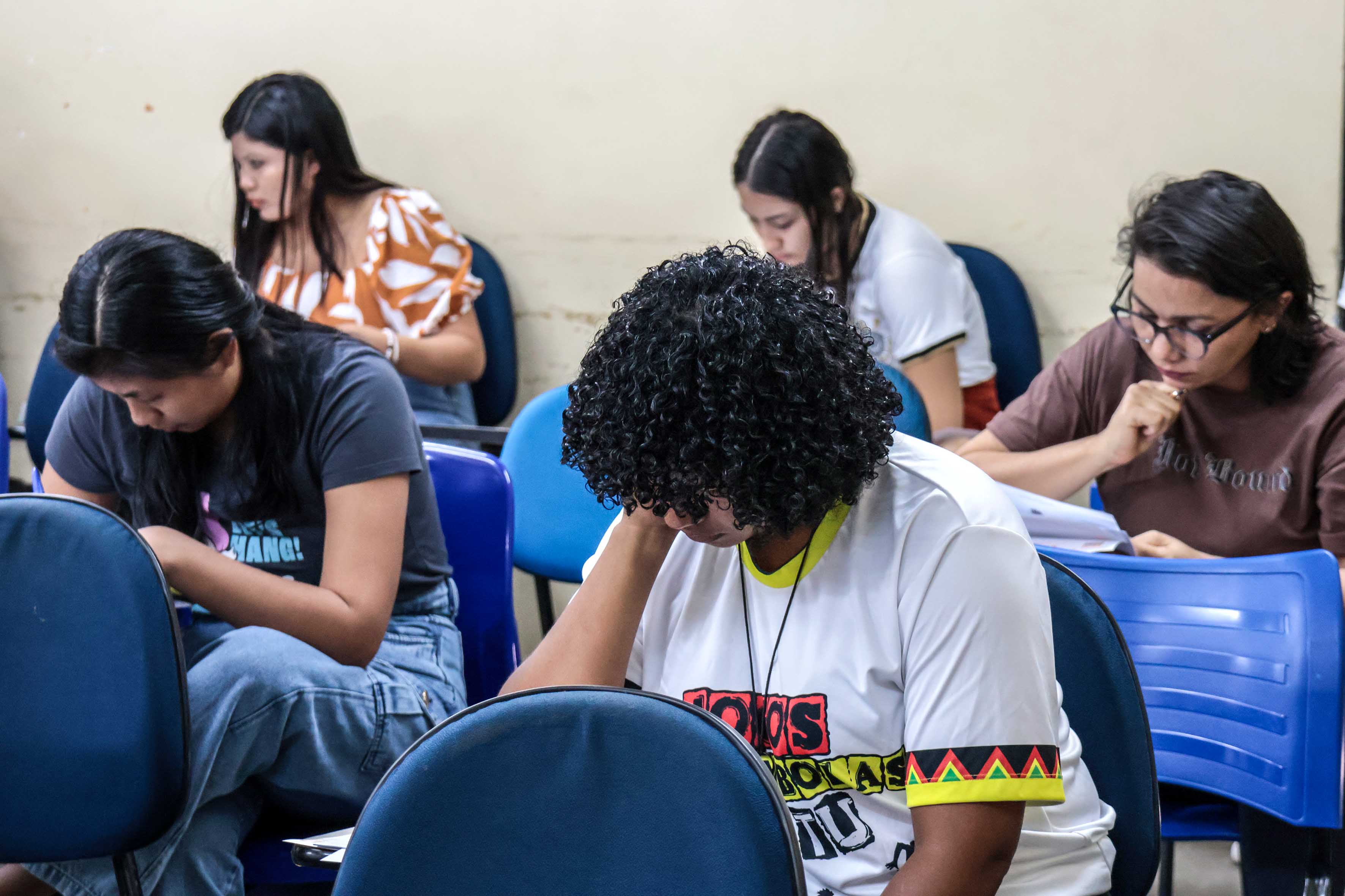 Seis pessoas estão sentadas em cadeiras escolares azuis, concentradas em responder a uma prova ou atividade escrita. Todas estão com a cabeça baixa, focadas em folhas de papel. Em primeiro plano, uma jovem de cabelo cacheado veste uma camiseta branca com a frase “Jovens Quilombolas e Indígenas”. As outras participantes, com diferentes estilos de cabelo e roupas, também estão atentas à tarefa. A sala é simples, com paredes claras e sinais de uso.