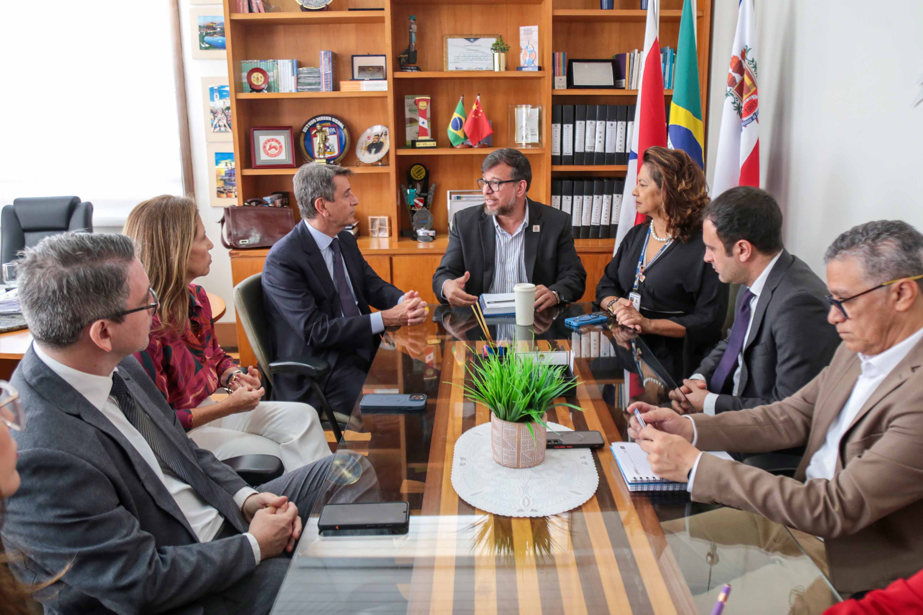  A imagem mostra um grupo de sete pessoas sentadas ao redor de uma mesa retangular em uma sala de reuniões bem iluminada e formal.  Na mesa, há uma pequena planta em vaso, vários documentos e alguns tablets digitais na frente de algumas pessoas. 