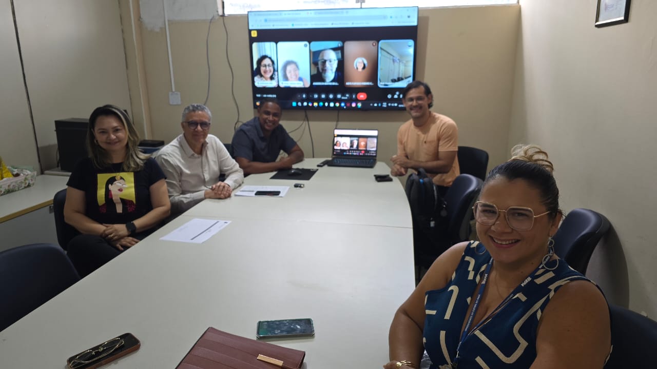 A imagem mostra cinco integrantes da comissão estão reunidos em uma sala, sentados ao redor de uma mesa, enquanto participam de uma videoconferência com outros membros exibidos em uma tela ao fundo. A imagem reflete um ambiente de trabalho colaborativo e híbrido.