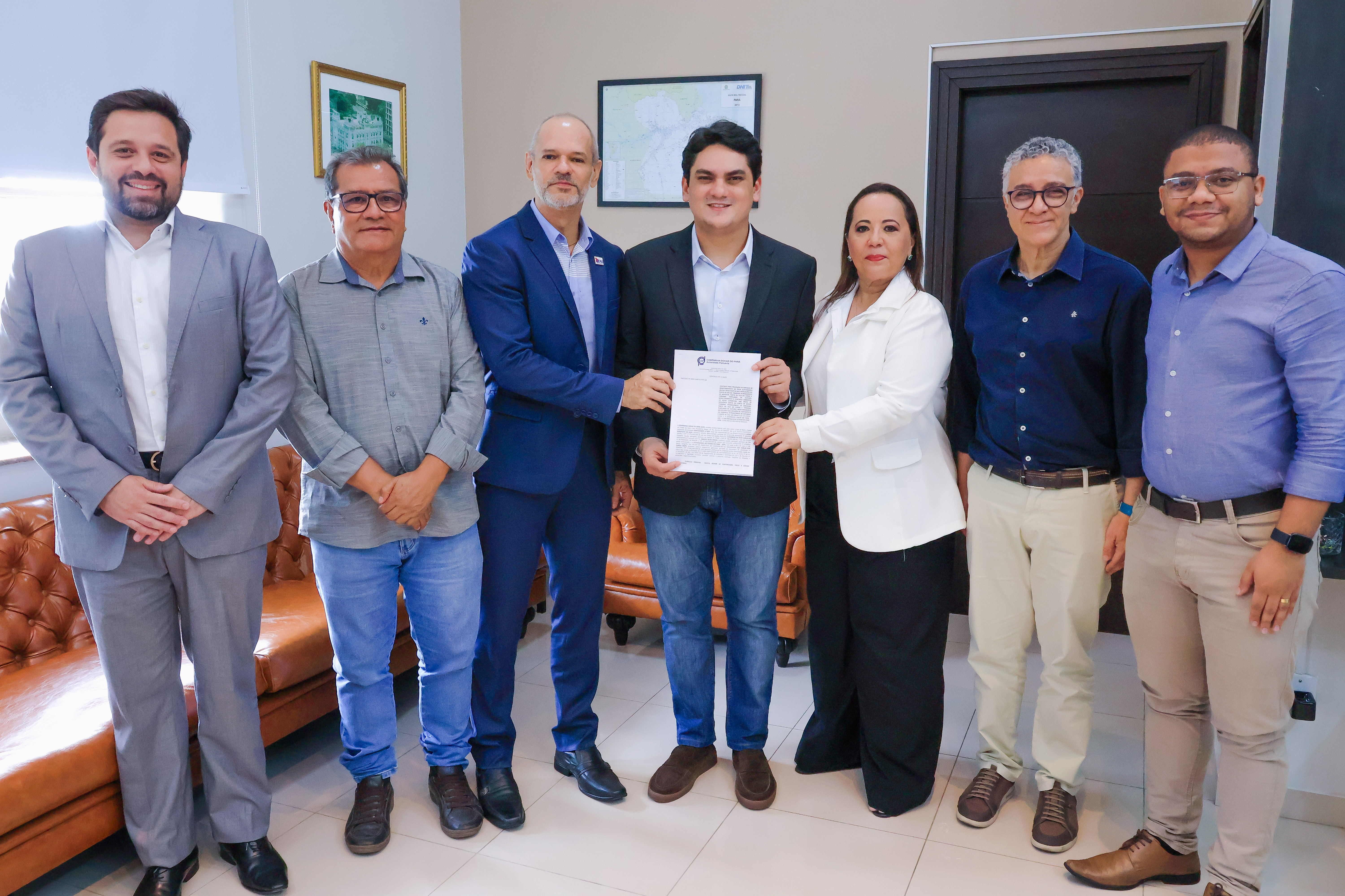 A imagem mostra sete pessoas posando em um ambiente corporativo, com sofás de couro e quadros na parede. O grupo é composto por homens e uma mulher, vestidos com trajes formais e casuais. No centro, um homem segura um documento, sendo acompanhado por outros dois que tocam o papel, sugerindo um momento de entrega ou assinatura oficial. Todos estão sorrindo, indicando um clima positivo e institucional.