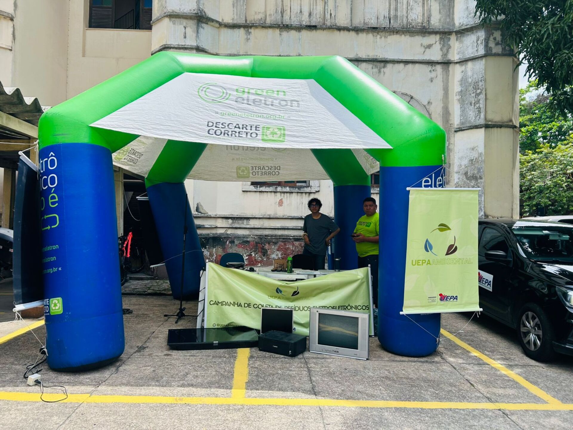 A imagem mostra uma tenda inflável grande nas cores verde e azul, montada em um estacionamento, para uma Campanha de Coleta de Resíduos Eletrônicos. No topo da tenda, há uma faixa com o logo “Green Eletron” e a mensagem “DESCARTE CORRETO”, e, abaixo, uma mesa com dois monitores antigos e outros equipamentos eletrônicos em desuso. Dois homens estão parados atrás da mesa, e no lado direito, há um banner vertical que exibe o logo da Uepa e um ícone de folha, indicando a natureza ecológica da iniciativa.