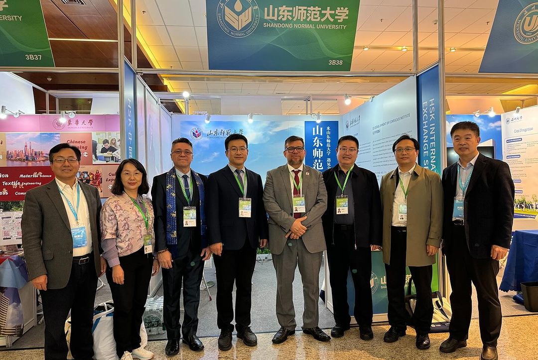 A imagem mostra um grupo de oito pessoas posando para uma foto em frente ao estande da Shandong Normal University em um evento. Todos estão formalmente vestidos, com crachás pendurados no pescoço, sugerindo participação em uma feira ou conferência acadêmica. O estande exibe banners promocionais com informações sobre a universidade e seus programas de intercâmbio internacional. Há elementos visuais relacionados à cultura e à educação chinesa nos materiais educaionais ao fundo.