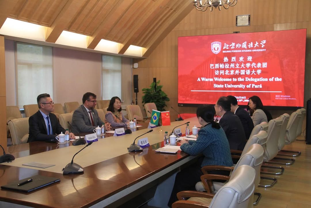 A imagem mostra uma reunião formal em uma sala de conferências. À esquerda da mesa, há representantes da Universidade do Estado do Pará. No centro da mesa estão as bandeiras do Brasil e da China, representando o tom diplomático e colaborativo do encontro. À direita, outros participantes estão engajados na conversa. Ao fundo, um telão exibe a mensagem: "A Warm Welcome to the Delegation of the State University of Pará", junto com o logotipo da Beijing Foreign Studies University e texto em chinês. 