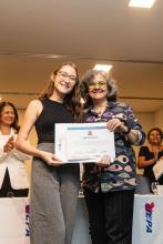 Aluna de óculos e calça cinza recebe certificado ao lado de uma mulher de óculos verdes, em clima de confraternização.