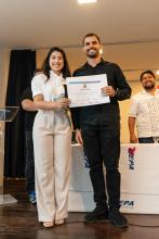 Aluno de camisa preta recebe certificado ao lado de uma mulher de branco, sorridente, durante a cerimônia.