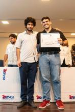 Aluno sorridente recebe o certificado no palco, posando ao lado de um colega e de um representante institucional.