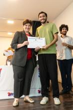 Aluno de camisa verde recebe certificado das mãos de uma autoridade feminina, ambos posando sorridentes.