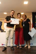 Entrega de certificado no palco. Um aluno recebe o documento e posa para foto ao lado de duas mulheres da organização.