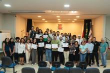 Grupo grande de alunos e equipe organizadora reunidos no auditório para foto oficial. Os formandos exibem seus certificados, enquanto professores e coordenadores acompanham. Ao fundo, bandeiras e o brasão da Uepa.